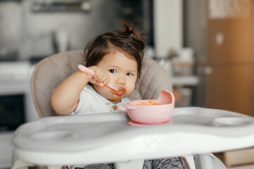 The child himself eats food at home in the kitchen. Cute little Asian girl eats fruit puree. The child is sitting in a high chair. The girl eats with a spoon. The child himself eats food