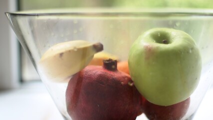 a mix of whole fruits refreshed with water lies in a large glass dish with the texture of water drops on a transparent surface on the kitchen table against the background of a summer window