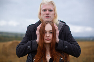 An interesting albino guy stands near a red-haired girl in the middle of a field and holds her head with his hands. Young couple in love. High quality photo