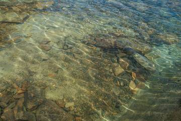 Meditative ripple of mountain lake. Beautiful relaxing background of stony bottom in transparent water of glacial lake in sunlight. Sunny backdrop with many stones in clear water of glacier lake.