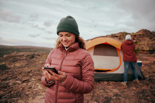 Happy young woman in winter clothing using smartphone during camping in mountain hill while man building tent in background