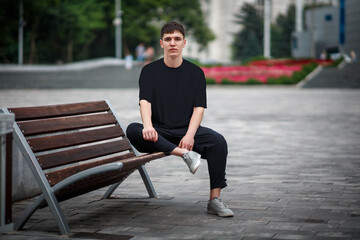Mockup of a black casual t-shirt on a guy sitting on a blurred background of a park, on a bench,...