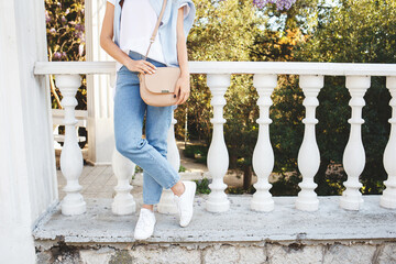 Bag in women's hands. Details of casual summer or spring outfit. Woman wearing blue jeans, white t shirt, sneakers, small beige cross body bag standing outdoors. Everyday look. Street fashion. No face