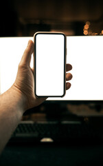 Mockup of hand holding modern smartphone with pc monitor on the background. Blank screen design template