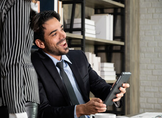 Fototapeta premium Two business people working together in office. Businessman sitting in office desk and using tablet while business woman standing next to him..
