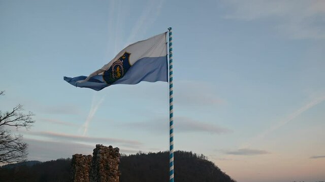 Flag of small town Kamnik waving in light breeze on top of ruins of old castle. Beautiful lights of sunset in Slovenia. Static shot, real time