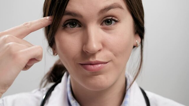 Doctor shows stupid. Close-up of irritated woman doctor on gray background looking at camera and twirls her finger at her temple. Slow motion