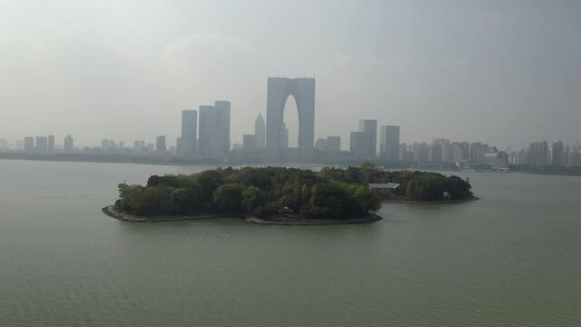 Aerial shot of island at Jinji Lake against buildings, drone ascending forward towards cityscape against sky - Suzhou, China