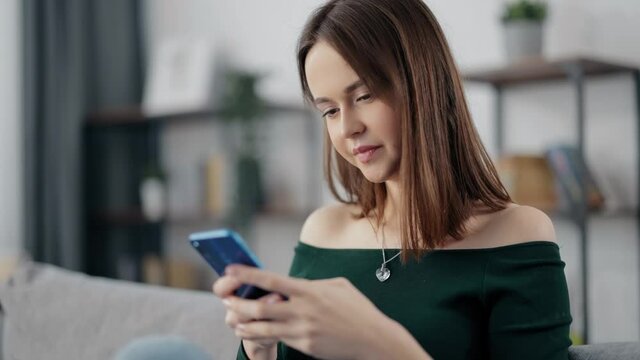 Beautiful happy woman relaxing on grey couch and using modern smartphone. Charming female with brown hair wearing stylish sweater with bare shoulders.