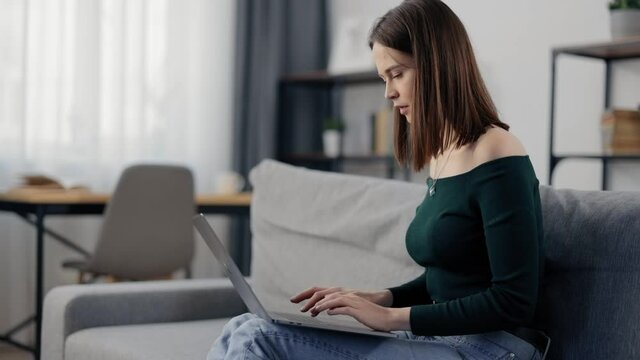 Stunning woman with brown hair browsing internet on wireless laptop while sitting on grey couch. Happy female using portable computer during free time at home.