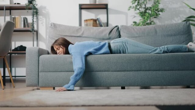 Tired young woman lying comfortably on grey couch at bright living room. Relaxation after hard long day. Cozy atmosphere at home.