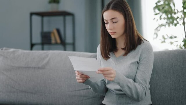 Young woman in casual wear looks upset while reading paper notification that holding in hands. Attractive brunette receiving bad news in letter.
