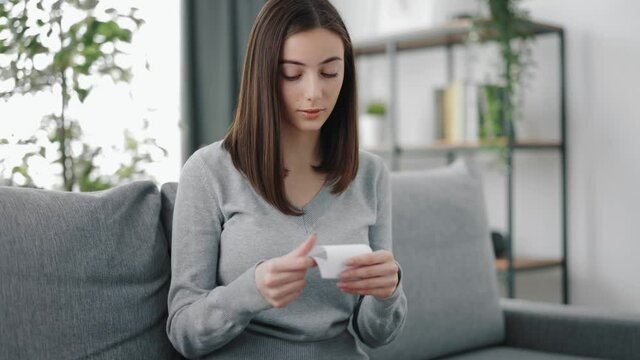 Smiling young woman with brown hair opening envelope and reading note with good news. Pretty female in casual outfit sitting on couch and holding letter.