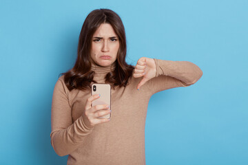 Fototapeta premium Displeased young brown haired woman casual beige turtleneck holds mobile phone, showing thumb down isolated on blue color background, frowning face and pout lips.