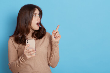 Young fun woman dressing casual beige turtleneck, holding mobile cell phone, pointing fingers aside on copy space isolated over blue background, amazed and surprised expression.