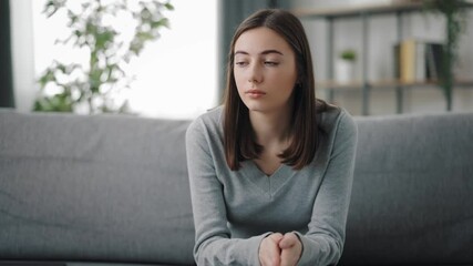 Pensive young woman in casual outfit sitting on grey couch and worried about something. Pretty brunette staying alone at home with bad thoughts. - Powered by Adobe