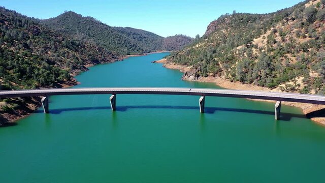 Aerial shot of bridge over famous reservoir against clear sky, drone flying backward from mountains on sunny day - Don Pedro Reservoir, California