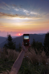 Arm of young man with a mobile phone on landscape background. Tourist taking picture of beautiful sunrise in mountain. Hands with smart phone. A man takes a photo for memory at dawn.