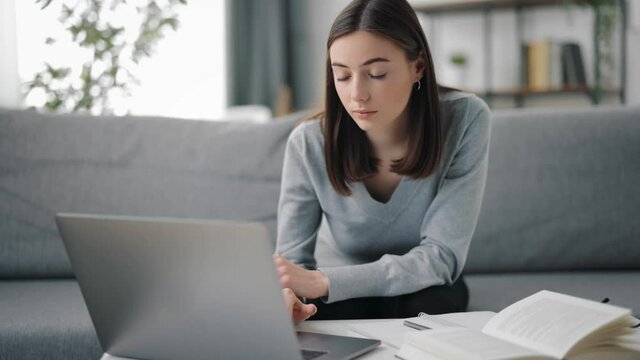 Pretty female student using portable laptop while studying during free time at home. Young woman with brown hair sitting at table with notes and books.