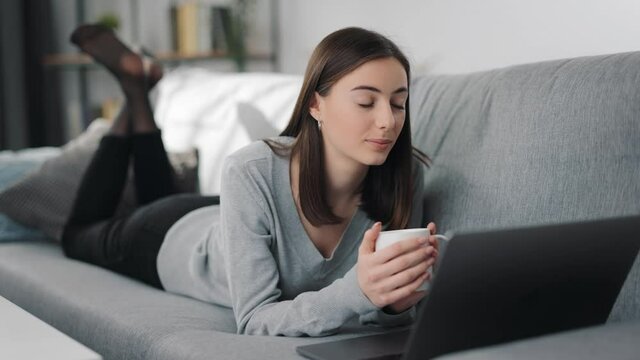 Relaxed young woman lying on couch with cup of tea in hands and looking at computer screen. Pretty lady with brown hair using modern technology during free time at home.