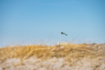 Fototapeta na wymiar Swedish flag fluttering on a pole, captured through the sandy dunes of Ribersborgsstranden (Ribersborg Strand or Beach). Scandinavian Swedish banderole waving on Ribersborgs Kallbadhus - Malmo, Sweden