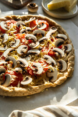 Homemade wholegrain pizza with cheddar cheese, mushrooms and tomatoes on baking paper on round kitchen table.