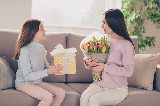 Profile photo of optimistic brunette red hairdo mom daughter hold flowers gift sit on sofa wear sweater pants at home