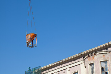 Fototapeta na wymiar Hanging orange concrete mixer against the blue sky and a fragment of a renovated building.