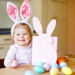 Obraz na płótnie Canvas Cute little toddler girl wearing Easter bunny ears playing with colored pastel eggs. Happy baby child unpacking gifts. Adorable healthy smiling kid in pink clothes enjoying family holiday