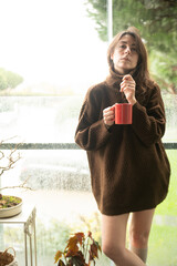 Portrait of sexy young woman holding a cup of coffee leaning against the window of her house, looking at the camera.