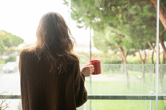 Woman Viewed From Behind Holding A Red Cup Of Coffee In Her Hand Looking Out The Window.Copy Space.Relax And Happy Time At Home