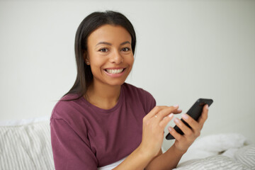 Charming young dark skinned woman with broad cute smile relaxing in bedroom enjoying online communication using wireless internet connection on mobile phone, messaging friend or reading inspiring post
