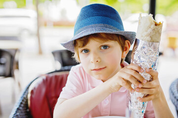 Cute healthy preschool kid boy eats turkish kebab wrap sitting in cafe outdoors. Happy child eating...