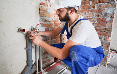 Horizontal snapshot of young plumber, wearing blue uniform and white helmet working with sealant fix of sewer pipe in kitchen or in bathroom in unfinished apartment