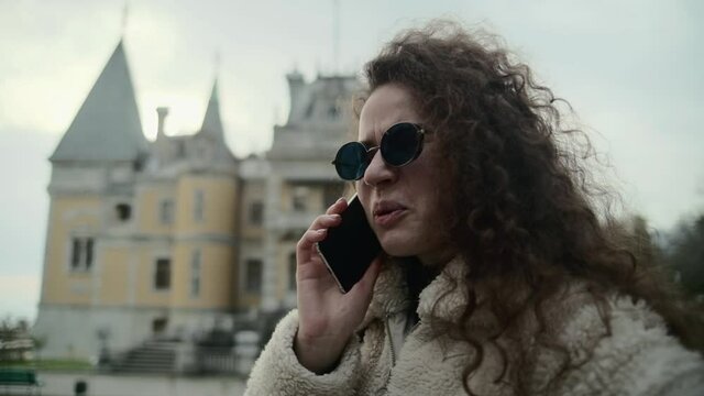 Displeased young businesswoman in sunglasses communicates on a mobile phone while standing in the street. An expensive building or palace is framed in the background. Roof with spiers. Girl laughs