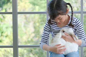A cute young girl take care and holding  a adorable white rabbit in the window background © Chavarin