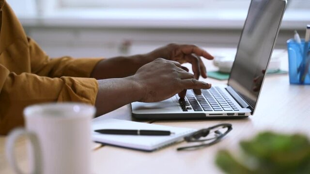 Businessman working with laptop and typing, sitting at table in home office spbas. Closeup view of young man types text on computer and does work, sits at desk in light interior. African American