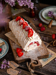 homemade tasty meringue roulade with white cream and strawberries on wooden board on rustic table with vintage green plates, forks, copper cups