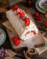 homemade tasty meringue roulade with white cream and strawberries on wooden board on rustic table with vintage green plates, forks, copper cups