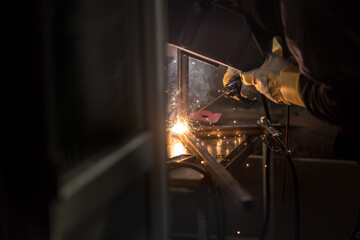 a welder in a welding mask, gloves welds metal with a welding machine and an electrode in his hand, sparks fly, a beautiful glow, bokeh, orange light on a black background, a flash of light and smoke