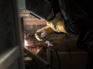 a welder in a welding mask, gloves welds metal with a welding machine and an electrode in his hand, sparks fly, a beautiful glow, bokeh, orange light on a black background, a flash of light and smoke