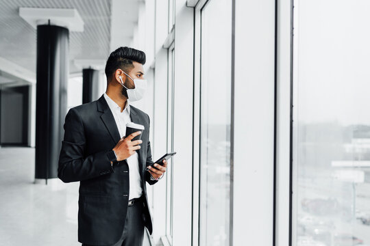 An Indian young businessman in a protective mask, in an office near a panoramic window, solves work problems with a phone in his hands and with coffee. Copy soace