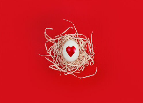 White egg with heart shape in decorative nest on red background.