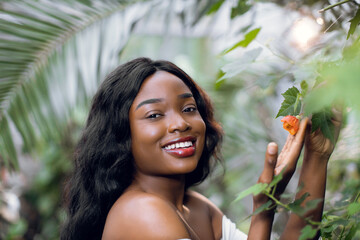 Pretty young afro american woman with perfect skin and natural make-up, posing to camera with smile, on the background of beautiful tropic plants, standing near flowering tree.