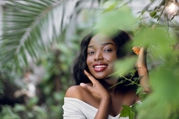 Beauty face. Woman model with natural makeup and healthy skin behind green plants. Portrait of beautiful black girl with nude makeup in tropical nature.