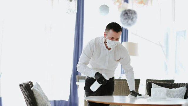 Male Waiter In Black Apron, Medical Mask And Gloves Cleaning White Table At The Restaurant By Disinfection Bottle. FHD