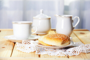 Fresh burger buns with sesame for breakfast at bright sunny morning. White chinaware cup, sugar...