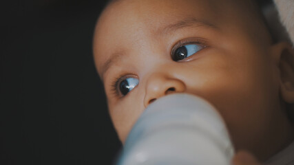 Adorable dark skin multiracial baby drinking his formula milk from the bottle in the hands of his mom. High quality photo