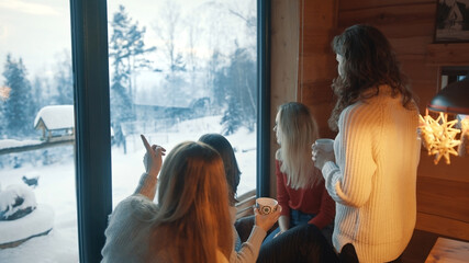Happy young women celebrating new year in wooden mountain hut. Relaxing near the window with hot...