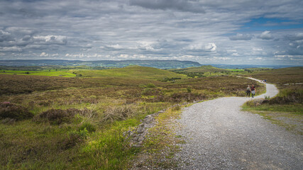 People walking on the trail between green hills and fields with stormy, dramatic sky in background, Cuilcagh Mountain Park, Northern Ireland